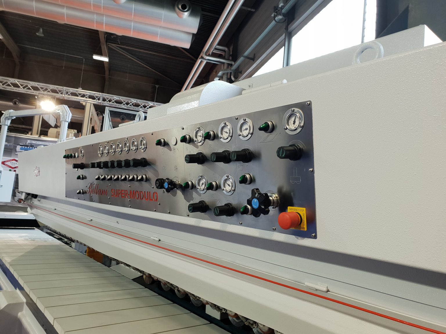 Grande plano do painel de controlo de uma grande máquina industrial, com indicadores, interruptores, botões e um botão vermelho de paragem de emergência, num salão de exposições bem iluminado da fábrica.