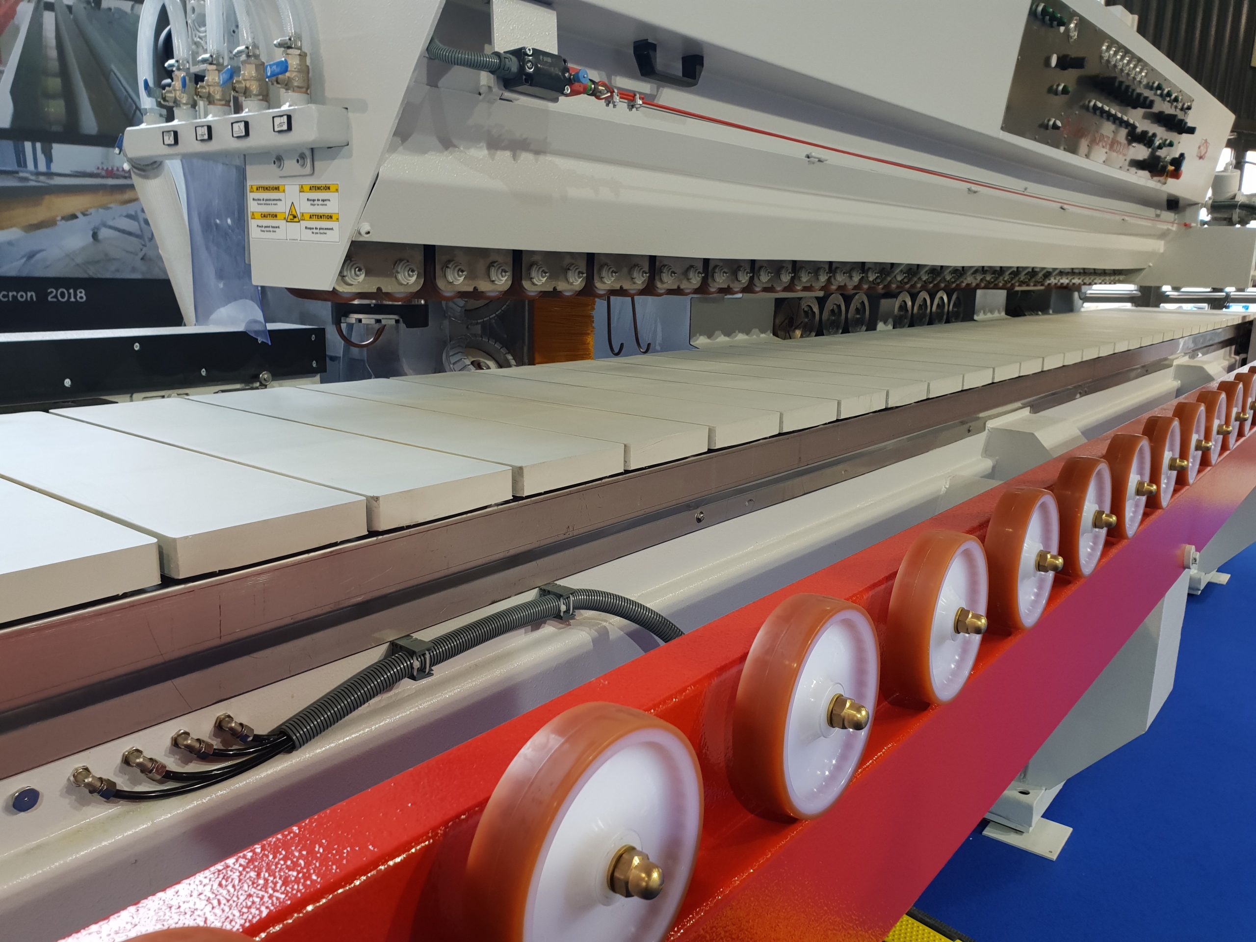 Máquina industrial de processamento de azulejos cerâmicos com filas de rolos, azulejos brancos e calhas metálicas de cor vermelha viva, num ambiente fabril organizado.