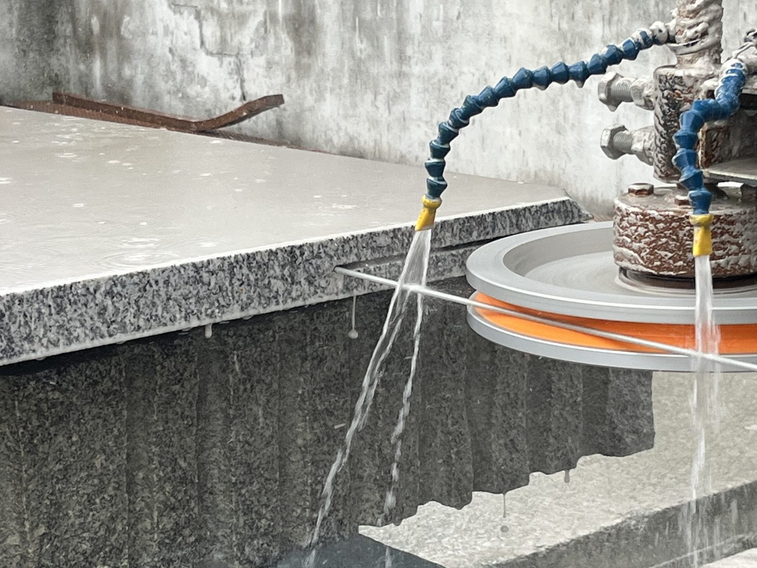 Mangueiras azuis pulverizam água sobre uma laje de granito ao lado de uma máquina de corte circular, criando um cenário industrial de corte de pedra com a superfície molhada.