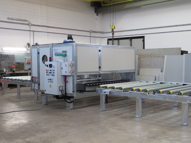 Piso de uma fábrica industrial com uma grande máquina de corte de metal, rolos e mesas transportadoras numa oficina limpa; é visível uma grua suspensa.