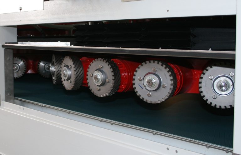 Grande plano de uma fila de discos industriais de esmerilagem ou lixagem montados num sistema transportador no interior de uma máquina de fabrico.