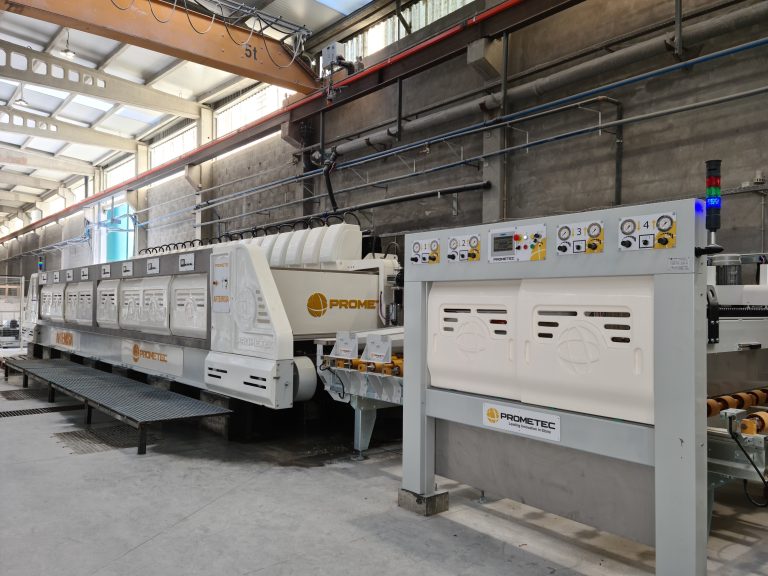 Máquina de fabrico têxtil industrial numa fábrica espaçosa, com painéis de controlo, rolos, vigas suspensas e luzes de trabalho intensas.