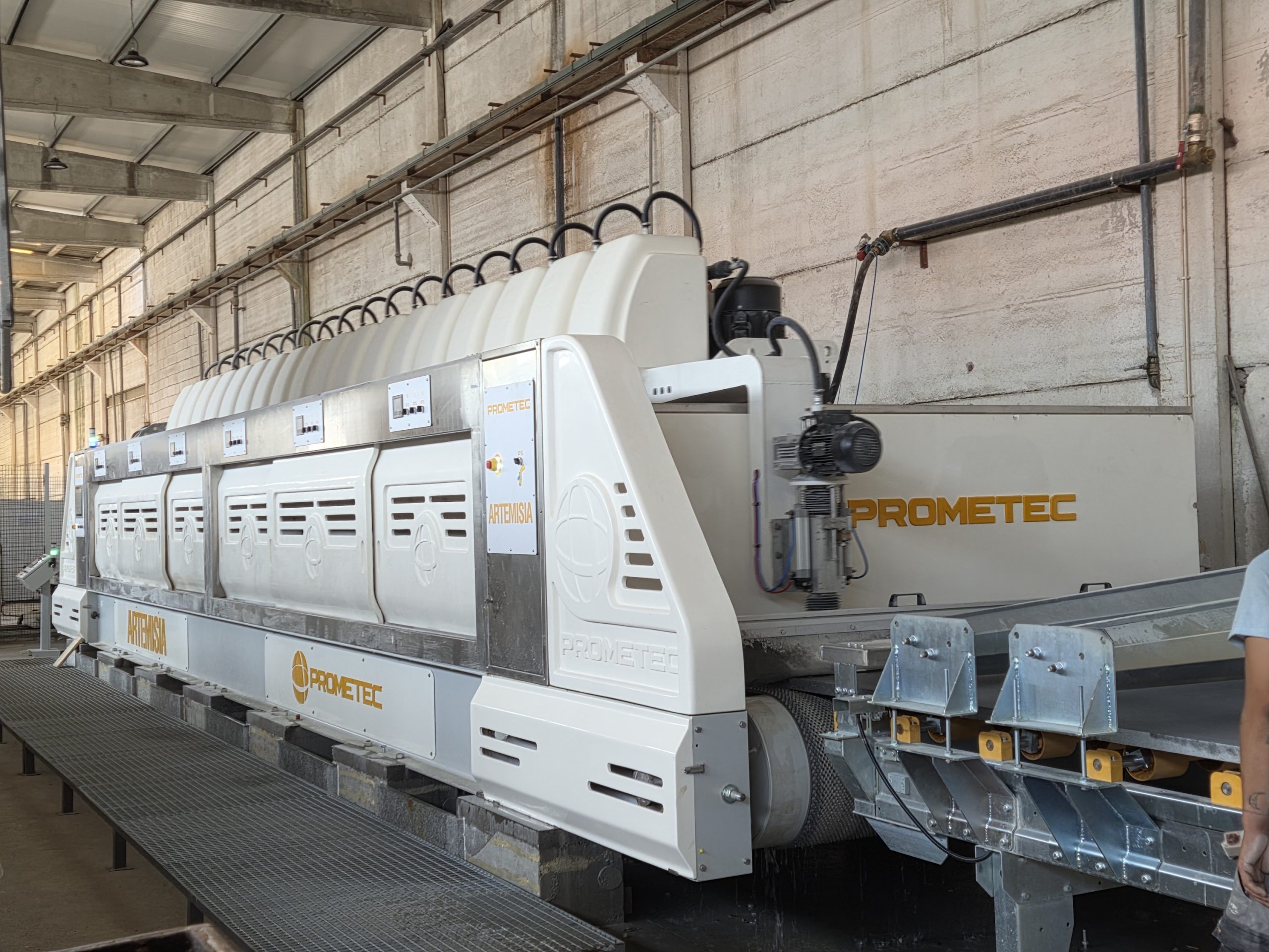 Máquina de processamento industrial Prometec num grande armazém, com estruturas metálicas, tubagens, peças de transportadores e iluminação superior intensa.