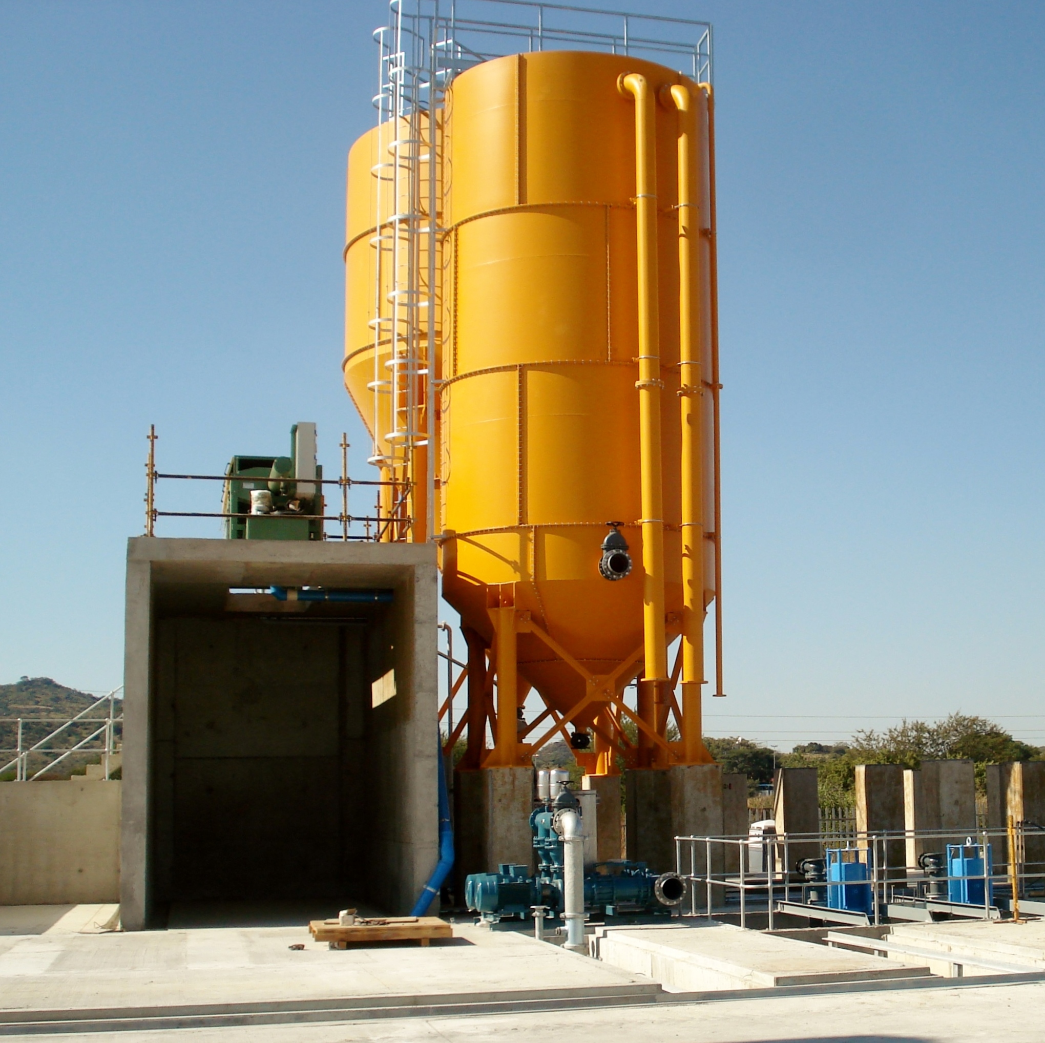 Grande silo industrial amarelo de armazenamento, com tubos, escadas e bombas, numa unidade de processamento de betão, sob um céu azul límpido, à luz do dia.