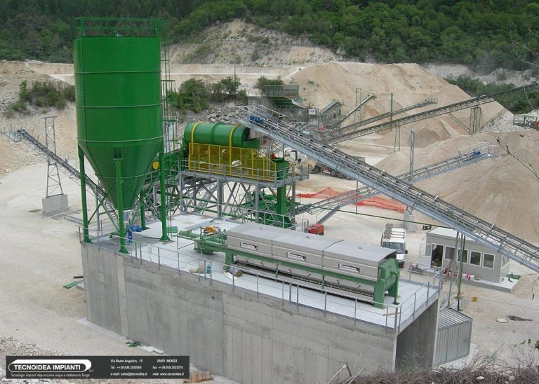 Instalação industrial de processamento de agregados numa pedreira, com tremonha verde, transportadores, equipamento de triagem numa plataforma de betão e colinas arborizadas.